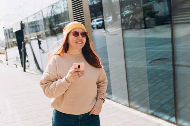 Sarı şapkalı ve pembe güneş gözlüklü 30 'lu yaşlardaki kız dışarıda akıllı telefon tutan sosyal medyayı kontrol ediyor. Şehir yaşam tarzı konsepti. Gezgin