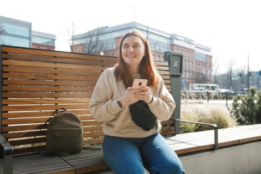 Açık havada akıllı telefon kullanan güzel, gülümseyen kız öğrenci. Şehir caddesinde bankta oturan neşeli, genç bir kadın. Şehir yaşam tarzı konsepti. Çevrimiçi sipariş ya da alışveriş kavramı