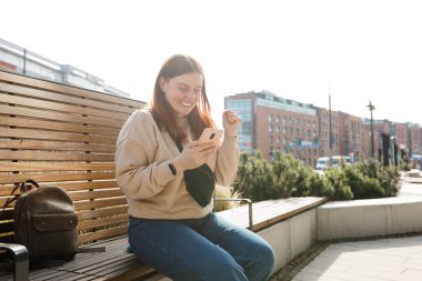 Cep telefonu tutan ve dışarıda kutlama yapan tatmin olmuş genç bir kadının portresi. Şehir caddesinde bankta oturan mutlu kız, hippi internet sitesinde online yarışmayı kazanmaktan çok memnun.