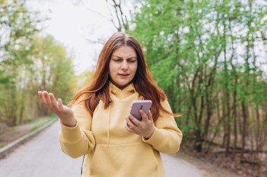 Şehir parkında dikilen üzgün kız. Negatif insan duyguları, şehir yaşam tarzı konsepti. Sırt çantalı üzgün genç bir kadın, baharda parkın dışında akıllı telefon kullanıyor..