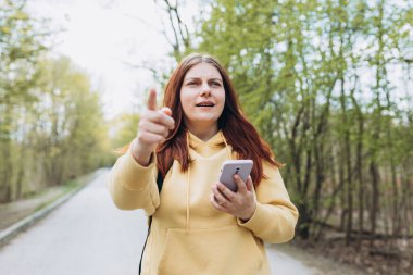 Şehir parkında dikilen üzgün kız. Negatif insan duyguları, şehir yaşam tarzı konsepti. Sırt çantalı üzgün genç bir kadın, baharda parkın dışında akıllı telefon kullanıyor. Gezgin