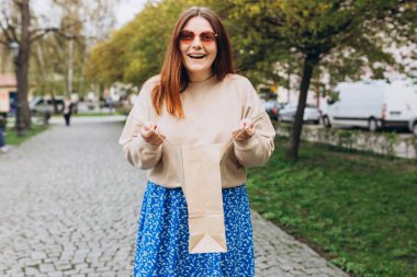 Güneş gözlüklü güzel kızıl kız dışarıda poz veriyor. Zarif, lüks bir kadının kese kâğıdı tutan açık hava moda portresi. Kentsel yaşam tarzı konsepti.