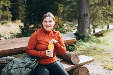 Yürüyüş kıyafetleri içinde kızıl saçlı mutlu bir kadın yürüyüş yaptıktan sonra duraklıyor. Su şişesinden su içen yürüyüşçü ya da termostan sıcak içecek.