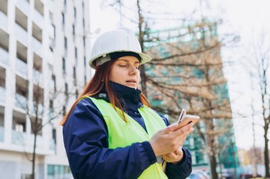 Açık havada akıllı telefon kullanan, dijital tabletli beyaz kasklı ve güvenlik yeleği olan genç mimar kadın. Kadın mühendis. Modern binanın yakınında lojistik, nakliye ve inşaat işçisi.