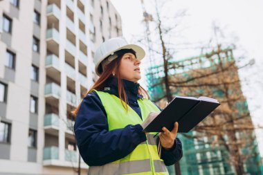 İnşaat sahasının yanında beyaz şapka ve yelek giyen kadın mühendis belgeleri, Mimari çek belgeleri ve modern bina yakınlarındaki iş akışı.
