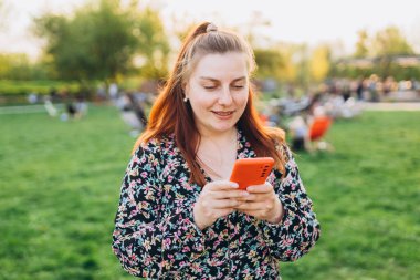 Doğa arka planında telefonla yürüyen gülümseyen güzel bir kadının portresi. Giyinik mutlu kadınlar, yaz mevsiminde parkta akıllı telefon kullanıyorlar. 30 'ların genç kızı kaygısızca dinleniyor.