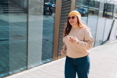 Sarı şapkalı ve pembe güneş gözlüklü 30 'lu yaşlardaki kız dışarıda akıllı telefon tutan sosyal medyayı kontrol ediyor. Şehir yaşam tarzı konsepti. Gezgin
