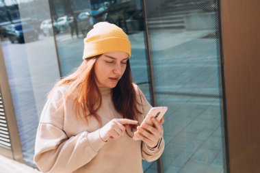 Şehir caddesinde yürüyen neşeli genç kadın akıllı telefonunu kontrol ediyor. Açık havada akıllı telefon kullanan sarı şapkalı güzel bir kadının portresi..