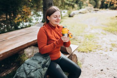 Yürüyüş kıyafetleri içinde kızıl saçlı mutlu bir kadın yürüyüş yaptıktan sonra duraklıyor. Su şişesinden su içen yürüyüşçü ya da termostan sıcak içecek.