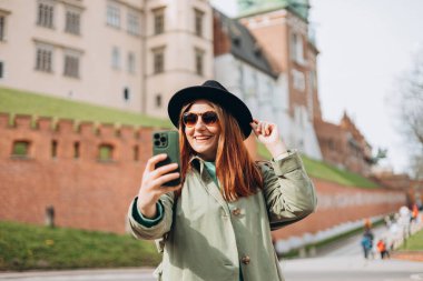 Çekici genç bayan turist yeni şehri keşfediyor. Krakow 'da elinde akıllı telefon tutan kızıl saçlı kız. Seyahat ediyorum. Şehirde yürüyen mutlu bir kız ve tarihi binaya karşı özçekim yapıyor.