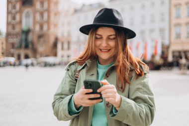 Bahar günü Krakow 'da Market Meydanı' nda yürüyen şapkalı güzel bir kadın ve elinde cep telefonu var. Şehir yaşam tarzı konsepti. Sosyal ağları kontrol edin, SMS ya da kitap oteli gönderin