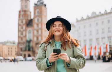 Bahar günü Krakow 'da Market Meydanı' nda yürüyen şapkalı güzel bir kadın ve elinde cep telefonu var. Şehir yaşam tarzı konsepti. Sosyal ağları kontrol edin, SMS ya da kitap oteli gönderin