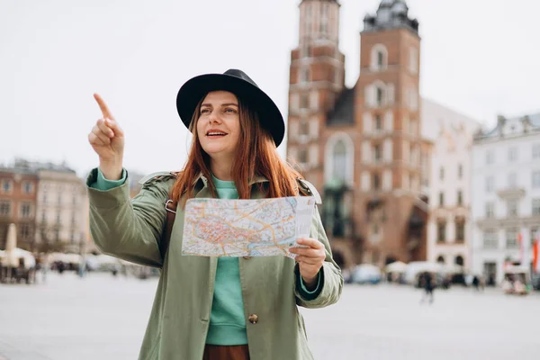 魅力的な女性観光客は新しい都市を探索しています 女性の指を指しているとクラクフのマーケット広場に紙の地図を保持再頭 秋にヨーロッパを旅行する セント メアリーズ大聖堂 — ストック写真