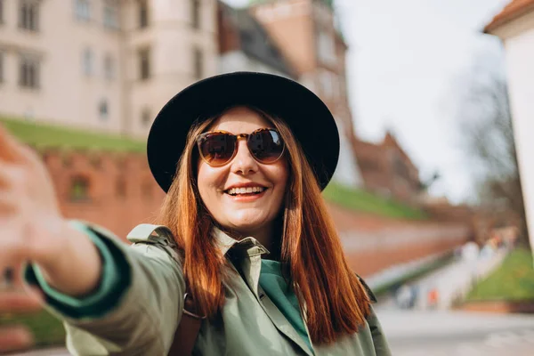 stock image Attractive young Redhead female tourist is exploring new city. Traveling. Happy optimistic girl walking in city and makes selfie poses against historic building in Krakow