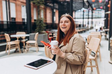 Genç bir kadın kahve içiyor, modern şehir caddesindeki kafe terasında oturuyor. Mutlu insan masada oturuyor ve açık havada akıllı telefon kullanıyor.