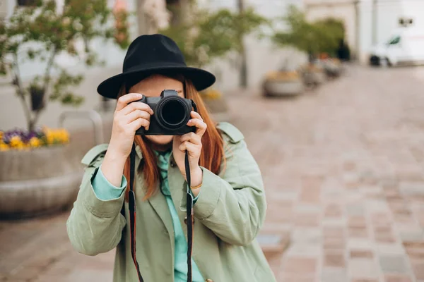 カメラで彼女の顔を覆う写真家の肖像 通りの背景にポーズバックパック付きのケアフリー女性 積極的な観光客屋外 陽気な30代の女の子で帽子あります探検都市 — ストック写真