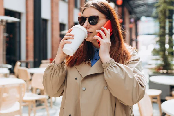 Moda ofis kıyafetleri içinde neşeli, genç, kızıl saçlı bir kadın ve telefonda bir fincan çayla konuşan güneş gözlüklü bir kadın. Mockup, elinde akıllı telefon ve biyo kahve fincanı tutan kadın. Kentsel yaşam tarzı konsepti