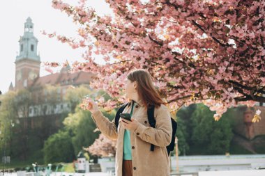 Bahar günü yürüyen ve arka planda cep telefonu tutan güzel bir kadın. Kızıl saçlı kız işaret ediyor. Telefon iletişimi. Şehir yaşam tarzı konsepti. Sosyal ağları kontrol et