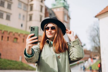 Çekici genç bayan turist yeni şehri keşfediyor. Krakow 'da elinde akıllı telefon tutan kızıl saçlı kız. Seyahat ediyorum. Şehirde yürüyen mutlu bir kız ve tarihi binaya karşı özçekim yapıyor.
