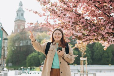 Bahar günü yürüyen ve arka planda cep telefonu tutan güzel bir kadın. Kızıl saçlı kız işaret ediyor. Telefon iletişimi. Şehir yaşam tarzı konsepti. Sosyal ağları kontrol et