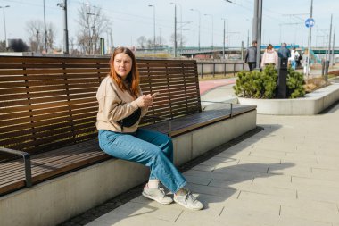 Açık havada akıllı telefon kullanan güzel, gülümseyen kız öğrenci. Şehir caddesinde bankta oturan neşeli, genç bir kadın. Şehir yaşam tarzı konsepti. Çevrimiçi sipariş ya da alışveriş kavramı