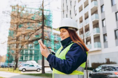 Açık havada akıllı telefon kullanan, dijital tabletli beyaz kasklı ve güvenlik yeleği olan genç mimar kadın. Kadın mühendis. Modern binanın yakınında lojistik, nakliye ve inşaat işçisi.
