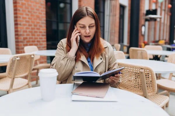 stock image Cheerful young redhead woman in fashion office clothes talking on Phone, 30s girl sitting on the cafe terrace on the modern city street. Female worker using on mobile Phone outdoors.