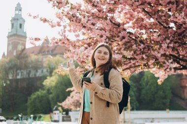 Bahar günü yürüyen ve arka planda cep telefonu tutan güzel bir kadın. Kızıl saçlı kız işaret ediyor. Telefon iletişimi. Şehir yaşam tarzı konsepti. Sosyal ağları kontrol et