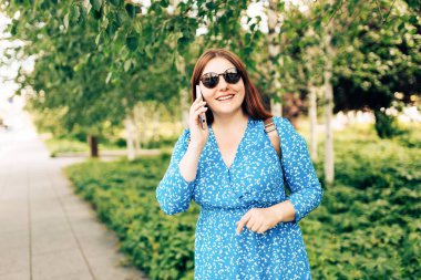 Güzel, gülümseyen bir bayan telefonda konuşuyor ve dışarıda duruyor. Telefon iletişimi. Şehir caddesinde yürüyen neşeli genç bir kadın, şehir yaşam tarzı konsepti..