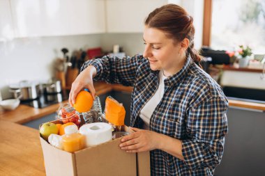 Genç kızıl saçlı kadın mutfakta içi taze organik yiyeceklerle dolu bir teslimat kutusu taşıyor. Evin arka planında farklı ürünlerden oluşan bir bağış kutusu. Ekspres teslimat, online alışveriş konsepti