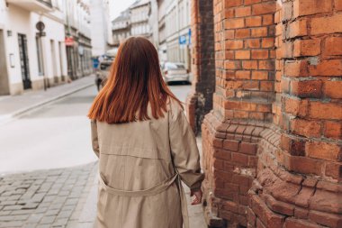 Uzun kızıl saçlı, Treanch giyen genç bir kadın şehir caddesinde yürüyor. Şehir arka planında yürüyen güzel bir kadın.