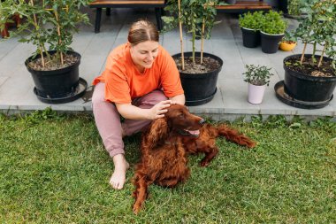 Yazın evin bahçesinde köpeğiyle oynayan neşeli, mutlu genç bir kadın. Güzel İrlandalı yerleşimci köpek çimenlerde yatıyor.