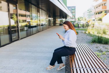 Ofisin yanındaki telefonu kullanarak gülümseyen iş kadını. Mavi gömlekli mutlu genç kadın akıllı telefonuna bakarak caddede yürüyor. Mobil cihaz üzerinde çalışan profesyonel yönetici