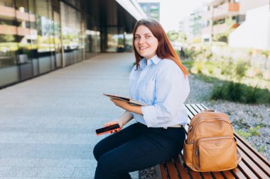 Caddede dokunmatik ped kullanan, kendine güvenen kızıl saçlı kadın yönetici, şehir arka planında bankta oturan kadınlar. Kentsel yaşam tarzı konsepti