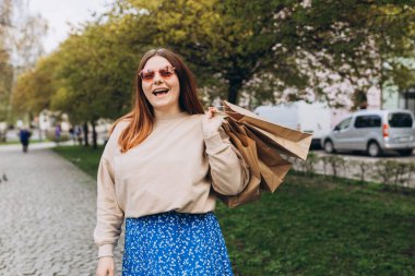 Elinde alışveriş poşetleri olan mutlu, genç, kızıl saçlı bir kadın, Kara Cuma 'da şehir caddesinde yürüyor. Şehir yaşam tarzı konsepti. Teslimat geliyor..