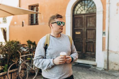 Sırt çantalı genç ve yakışıklı bir adam eski kasabanın arka planında kahve fincanı içiyor. Eski moda kafe terasında bir yaz günü. Çay içerek..