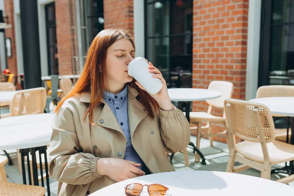 Moda ofis kıyafetleri içinde neşeli genç bir kadın ve eko-çay eşliğinde güneş gözlüğü. Mockup, elinde biyo-kahve fincanı tutan kadın ve caddedeki kafe terasında oturan kadın..