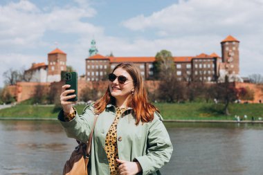Çekici genç bayan turist yeni şehri keşfediyor. Krakow 'da elinde akıllı telefon tutan kızıl saçlı kız. Seyahat ediyorum. Mutlu iyimser kız şehirde yürüyor ve tarihi binaya karşı özçekim yapıyor.