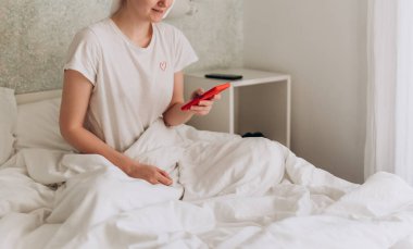 Telefonu olan genç bir kadın rahat bir yatakta yatıyor. Beyaz, 30 'lu yaşlarda bir kız evde yatarken akıllı telefon kullanıyor.