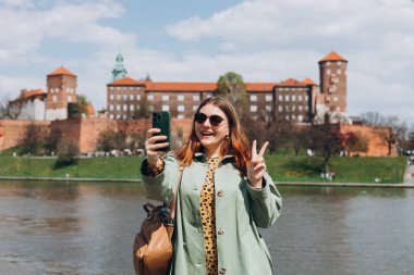 Çekici genç bayan turist yeni şehri keşfediyor. Krakow 'da elinde akıllı telefon tutan kızıl saçlı kız. Seyahat ediyorum. Mutlu iyimser kız şehirde yürüyor ve tarihi binaya karşı özçekim yapıyor.