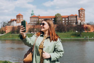 Çekici genç bayan turist yeni şehri keşfediyor. Krakow 'da elinde akıllı telefon tutan kızıl saçlı kız. Seyahat ediyorum. Mutlu iyimser kız şehirde yürüyor ve tarihi binaya karşı özçekim yapıyor.