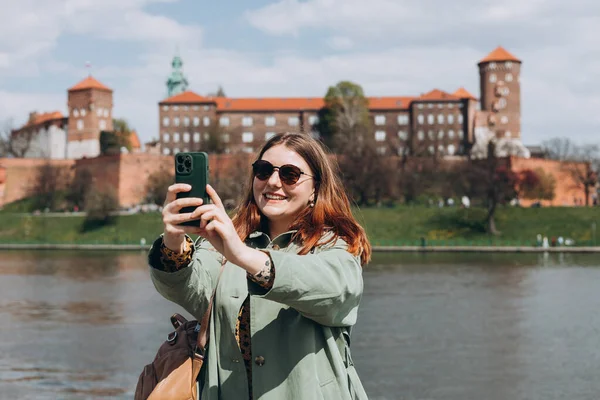 stock image Attractive young female tourist is exploring new city. Redhead girl holding a smartphone in Krakow. Traveling. Happy optimistic girl walking in city and makes selfie poses against historic building