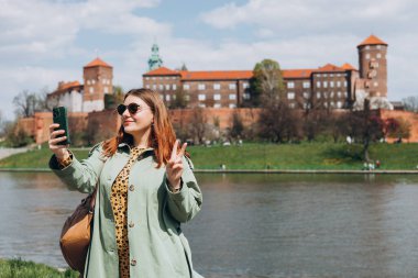 Çekici genç bayan turist yeni şehri keşfediyor. Krakow 'da elinde akıllı telefon tutan kızıl saçlı kız. Seyahat ediyorum. Mutlu iyimser kız şehirde yürüyor ve tarihi binaya karşı özçekim yapıyor.