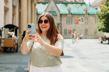 Güzel bir tatilde eski şehirde yürürken selfie çeken genç bir kızın yakın çekimi. Genç ve güzel bir kadın Bratislava 'nın tarihi bölümünde market sokağının önünde eğleniyor.