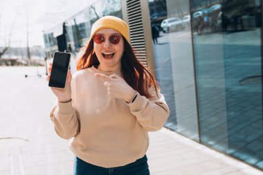 Kızıl saçlı bir kadının, boş siyah ekranlı akıllı bir telefonu işaret ederken ki görüntüsü. Şapkalı ve güneş gözlüklü heyecanlı bir kızın portresi cep telefonunu işaret ediyor.