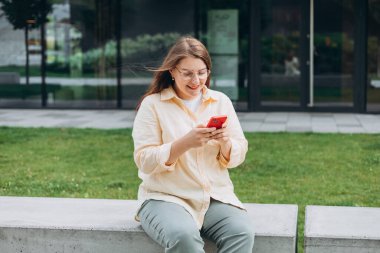Güneşli bir günde gülümseyen kadın akıllı telefondan mesaj atıyor. Şehir yaşam tarzı konsepti. Gezgin oturuyor ve akıllı telefondan haberleri okuyor, hızlı bağlantı, açık havada mobil uygulamaları kontrol ediyor..