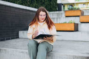 30 'lu yaşlardaki zarif bir kadın resmi olarak giyinmiş, dışarıda duruyor ve elinde belgelerle bir klasör tutuyor.