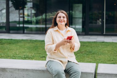 Güneşli bir günde gülümseyen kadın akıllı telefondan mesaj atıyor. Şehir yaşam tarzı konsepti. Gezgin oturuyor ve akıllı telefondan haberleri okuyor, hızlı bağlantı, açık havada mobil uygulamaları kontrol ediyor..