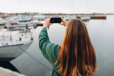 Liman telefonunda fotoğraf çeken bir kadın turist. Rıhtım boyunca yürüyen bir turist, deniz kenarında. Seyahat etmek için ilginç yerleri keşfederek tatil kavramı