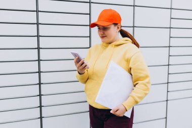 Genç bir kadın, elinde telefon ve küçük bir kutuyla duran kuryeden otomatik posta kutusundan bir paket aldı. Otomatik dolaplara hızlı teslimat kavramı